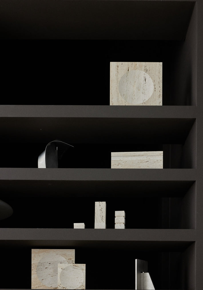 Medium tray | travertine displayed as a square stone block with a round indent, surrounded by minimalist geometric stone and metal objects on a matte-dark shelving unit.