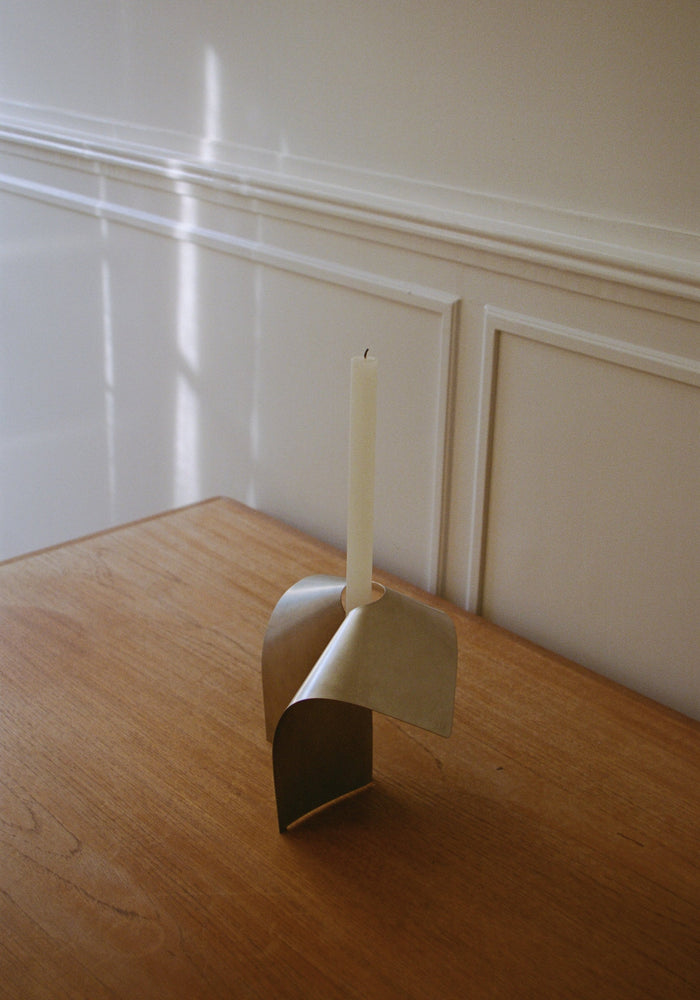 Stainless steel candle holder with a minimalist bent shape, featuring a brass-like finish, holding a white taper candle on a wooden tabletop. Handcrafted design by Paul Coenen.