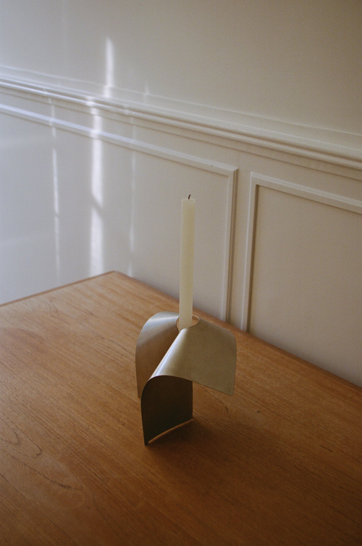 Minimalist modern stainless steel candle holder with a single white taper candle placed on a wooden table against a light-colored paneled wall, Stainless steel candle holder | Stainless steel.