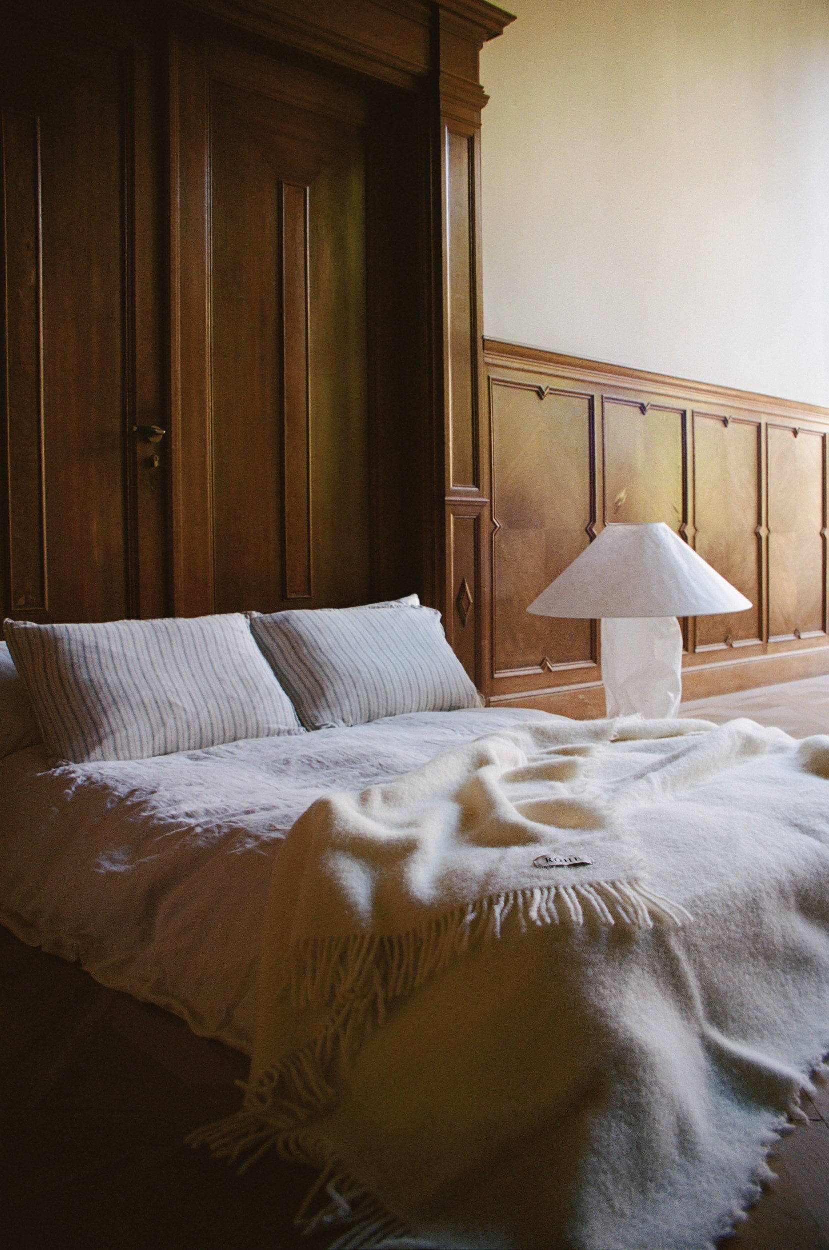 Bedroom with wooden paneling, bed with white bedding and wool fringed cream blanket and lamp on nightstand.