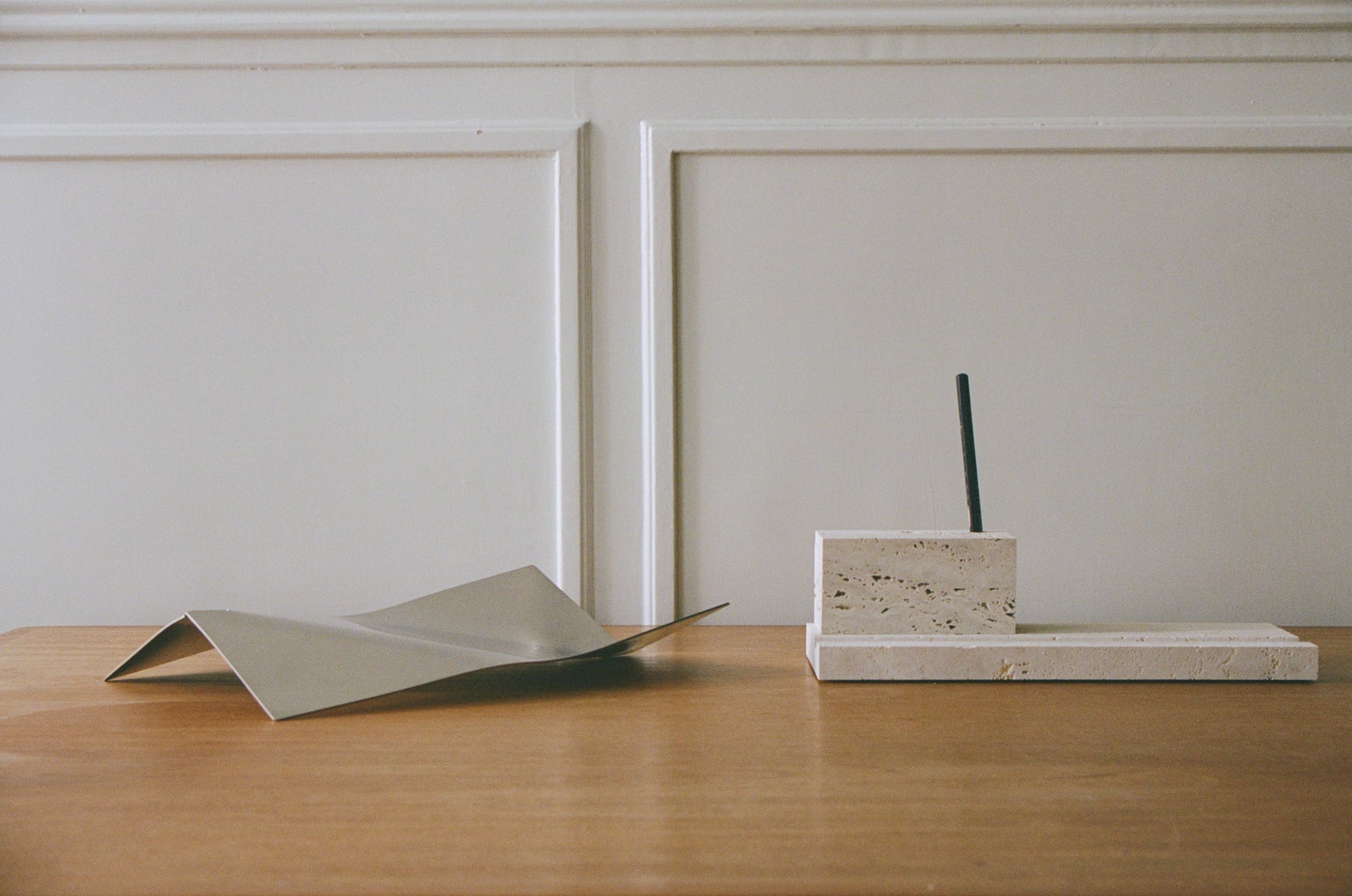 Wooden table with a stainless steel tray and travertin pencil holder placed on top against a white paneled wall