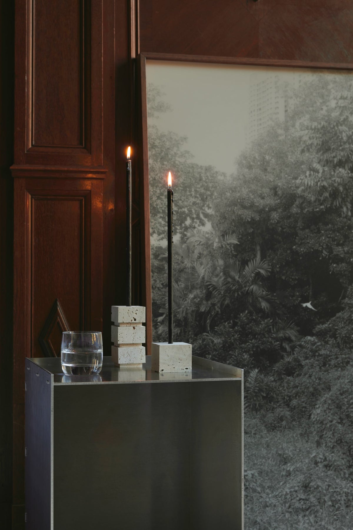 Sculptural candle holder small | travertine, featuring handcrafted stacked cubes and a single cube, holding two lit black taper candles on a metal pedestal.