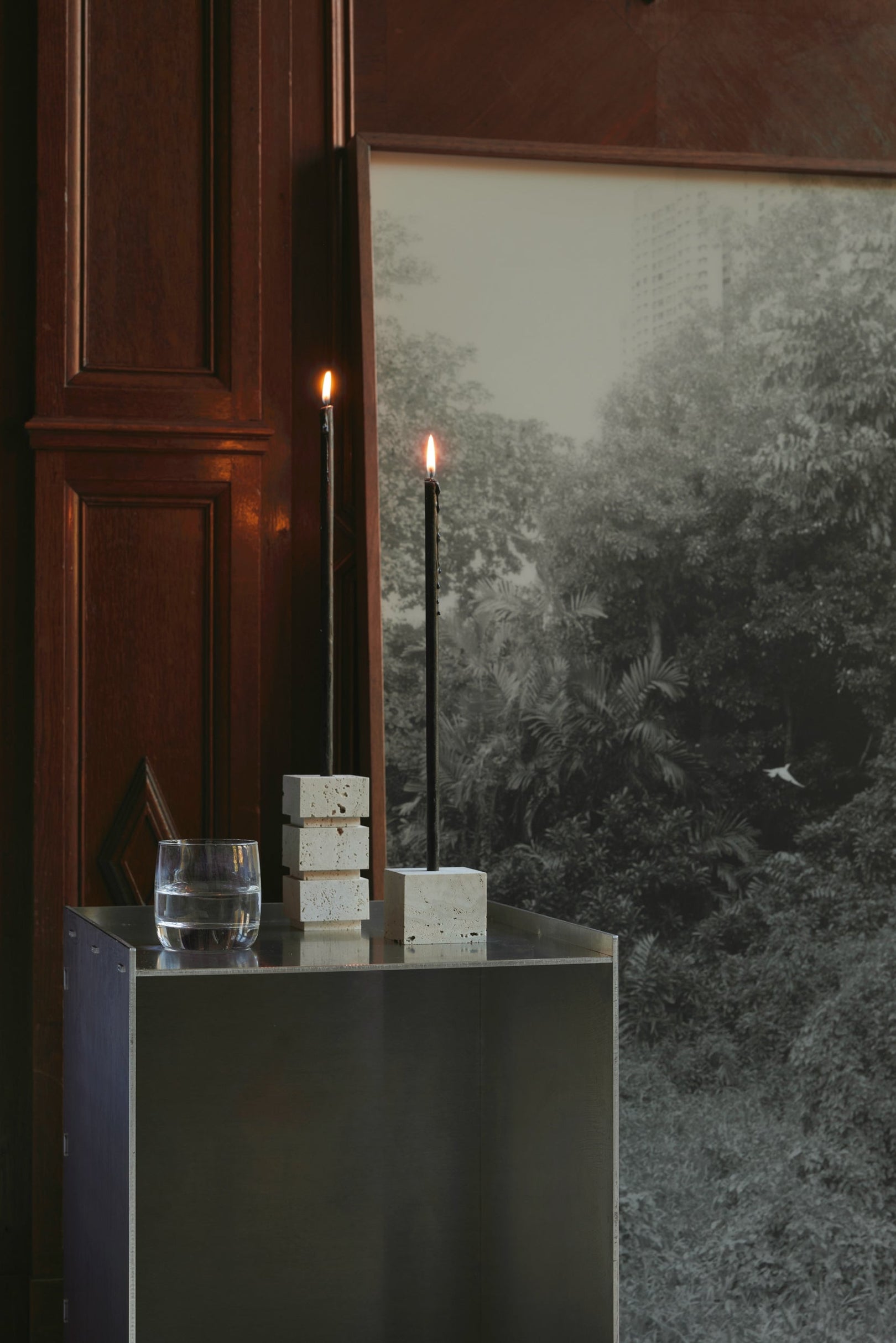 Sculptural candle holder small | travertine, featuring handcrafted stacked cubes and a single cube, holding two lit black taper candles on a metal pedestal.