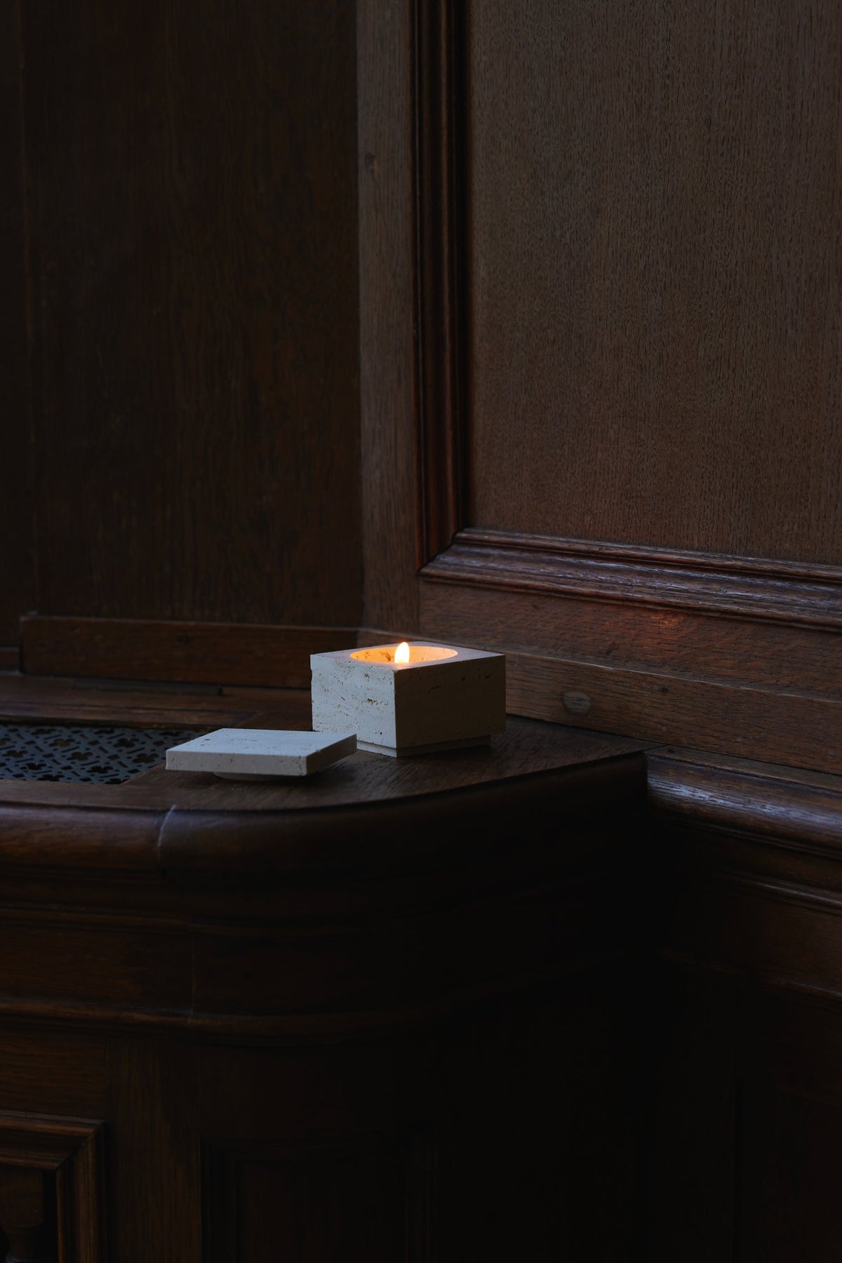 Tealight box | travertine: A lit candle in a square travertine holder with lid beside it on dark wood, highlighting its handcrafted Dutch design.