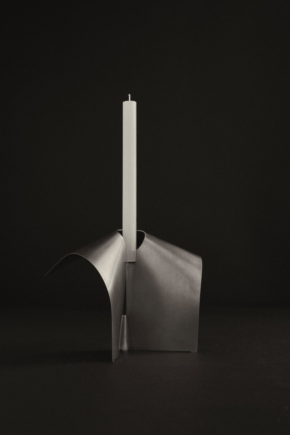 Stainless steel sculptural candle holder with a white dinner candle inside placed on a dark background