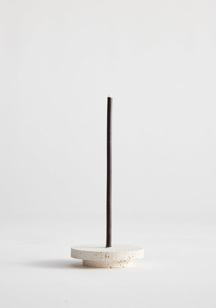Dish candleholder small | travertine: A black taper candle stands in a handcrafted, round travertine base, showcasing unique porous texture, crafted by artist Louis Crombag.