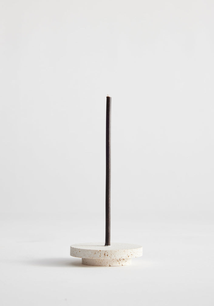 Dish candleholder small | travertine: A black taper candle stands in a handcrafted, round travertine base, showcasing unique porous texture, crafted by artist Louis Crombag.