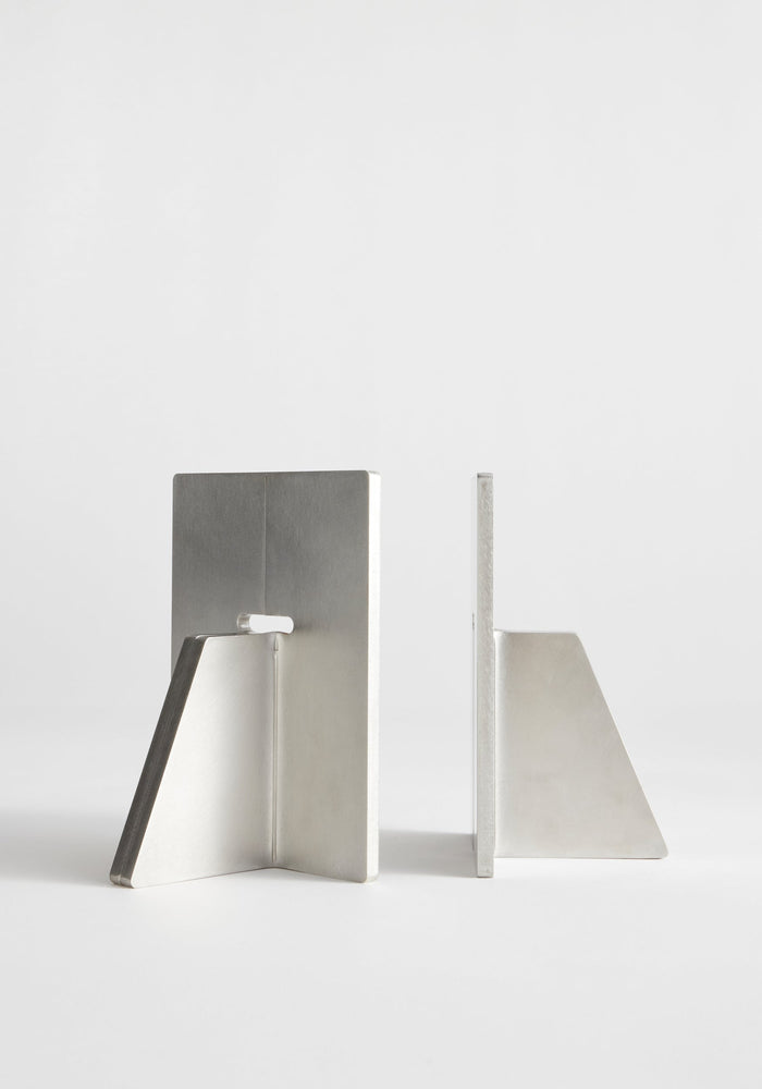 Stainless steel bookends: two brushed-aluminum brackets with a vertical rectangular plate and triangular base, handcrafted, showcasing one front view and one side profile on a white background.