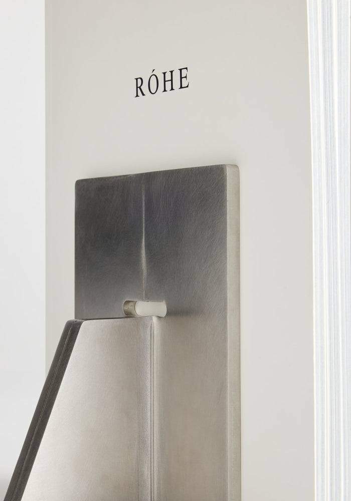 Stainless steel bookends: A close-up of a thick, white hardcover book titled “RÓHE” supported by one of two sculptural, handcrafted stainless-steel bookends.