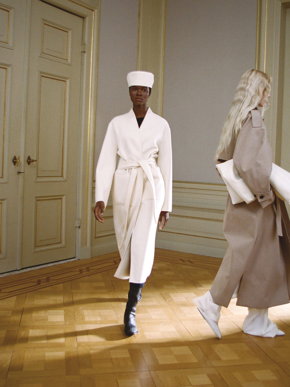 Two models walking down a runway, one dressed in a white belted coat, one in a taupe trenchcoat, in a room with wooden floors and paneled walls