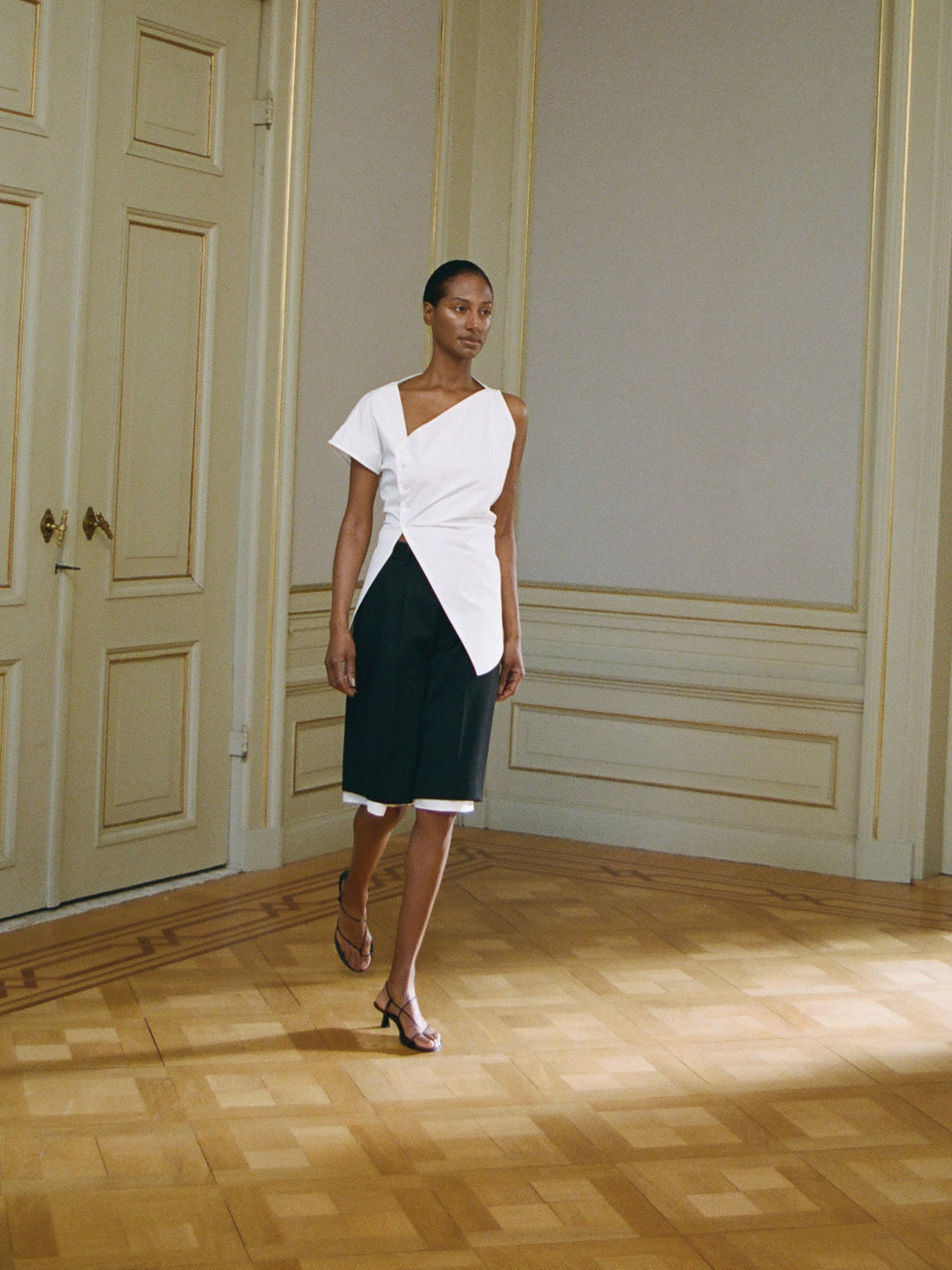 Woman in a white  asymmetrical poplin top and black shorts walking in a room with wooden flooring and paneled walls.
