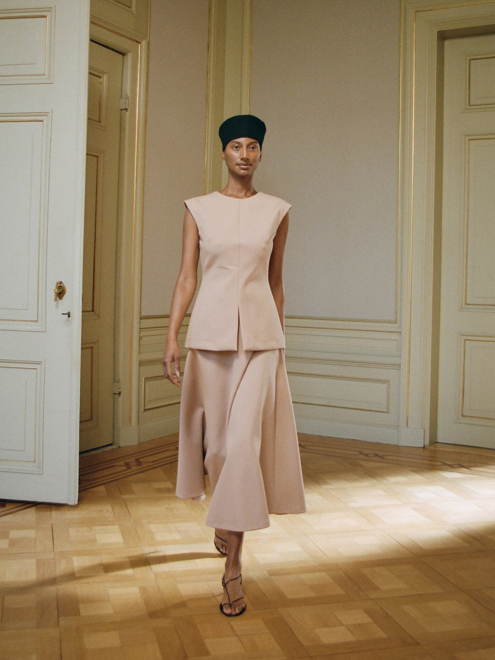 Woman in a pink powder colored top and matching a-line skirt standing in an elegant room with wooden floors and paneled walls