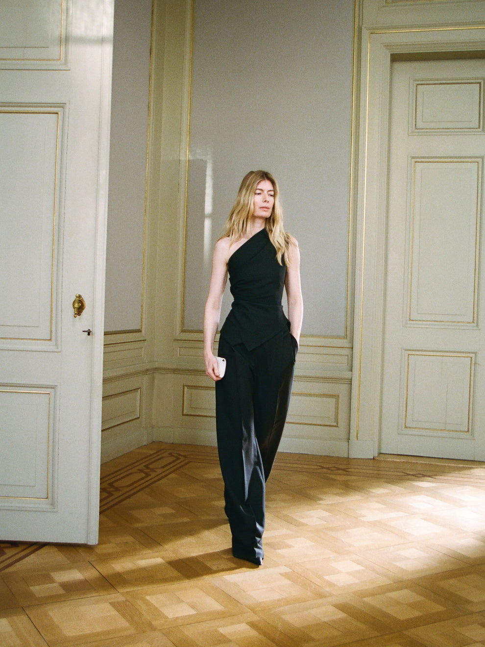 Woman in a black off shoulder top and black trousers standing in a room with paneled walls and wooden floors.