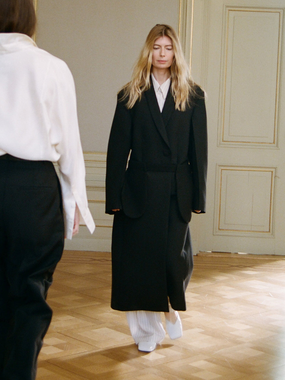 Woman in a black coat standing in a room with wooden floor and paneled walls