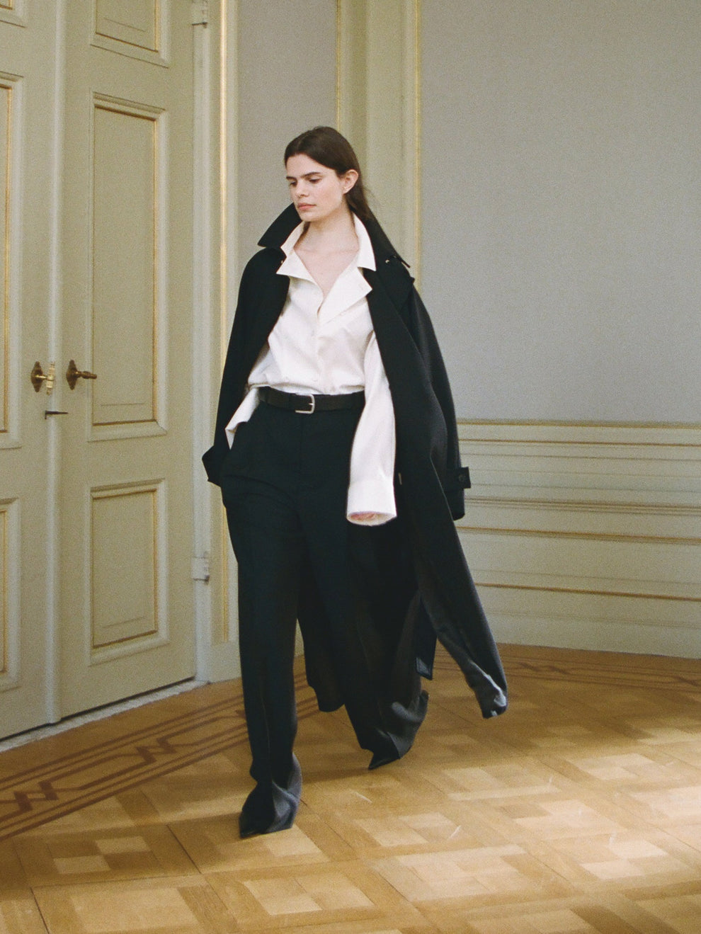 Woman in a black trenchcoat and white shirt, black trousers with a belt walking in a room with wooden flooring and a panelled door.