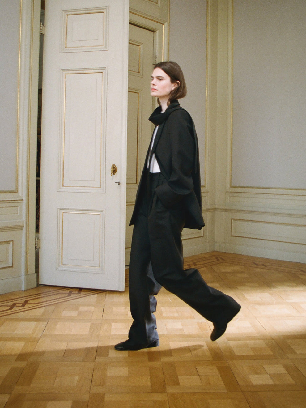 Woman walking in black suit in a room with wooden flooring and white walls