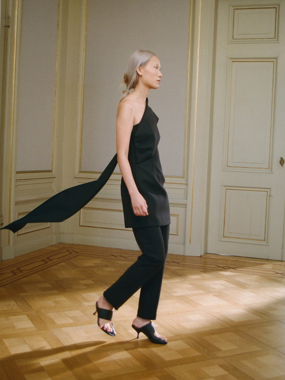 Woman in a black short dress with cape detail walking in a room with wooden floor and paneled walls