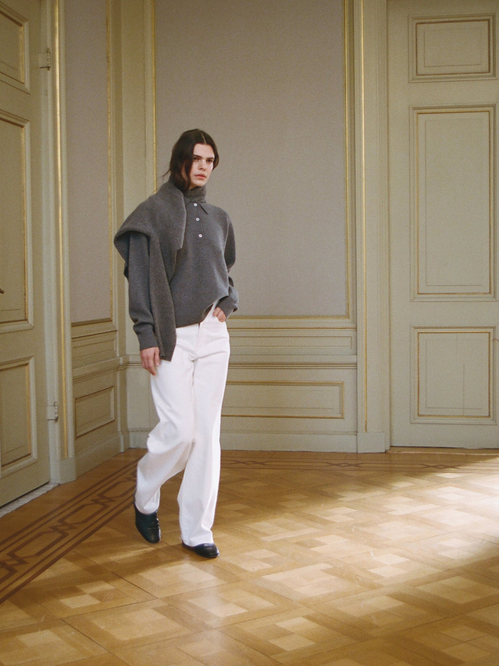 Woman wearing a gray polo sweater and white wide led jeans walking in a room with wooden flooring and paneled walls.