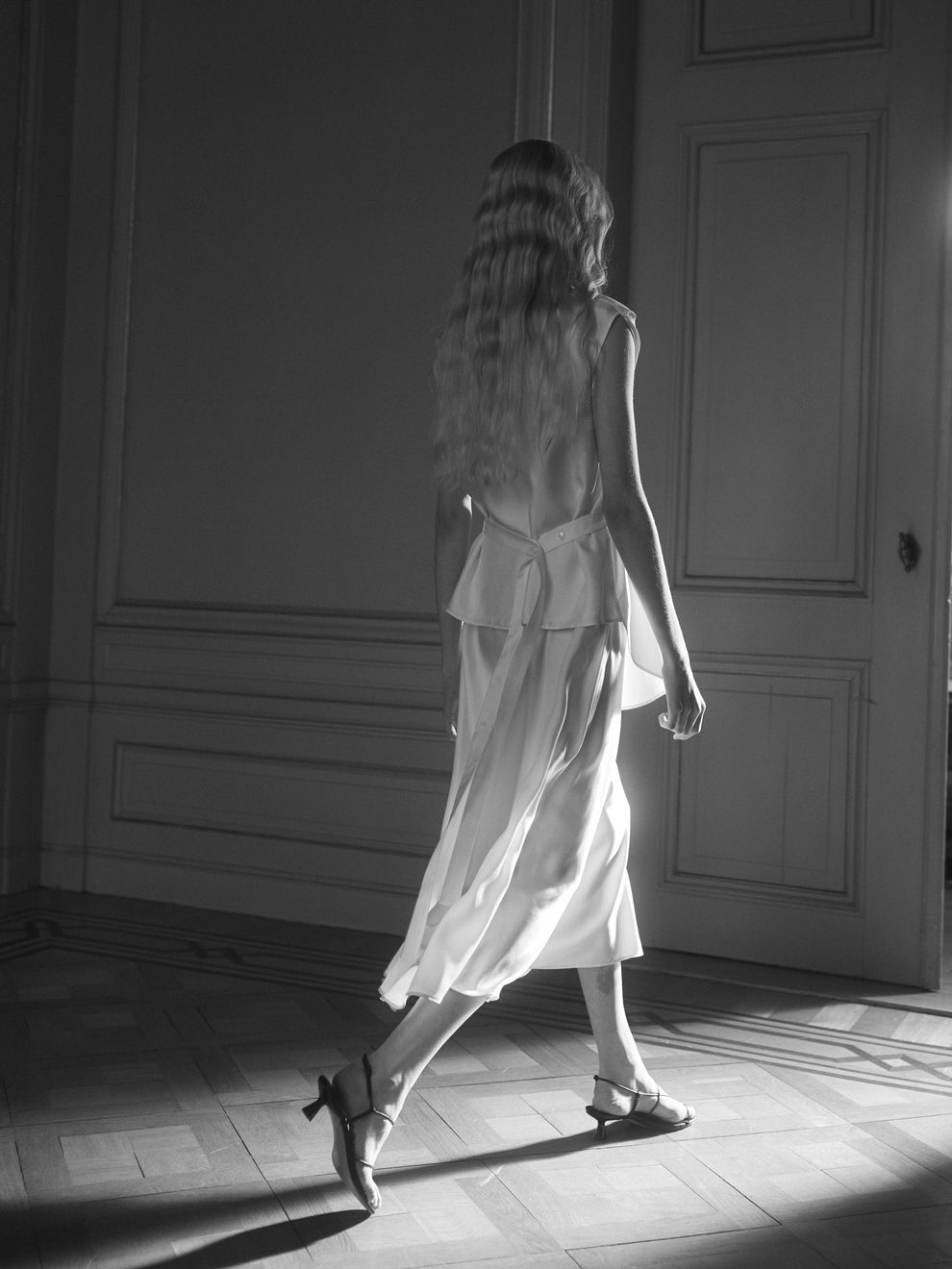 Woman walking away from a door in a dimly lit room in a silk flowy dress