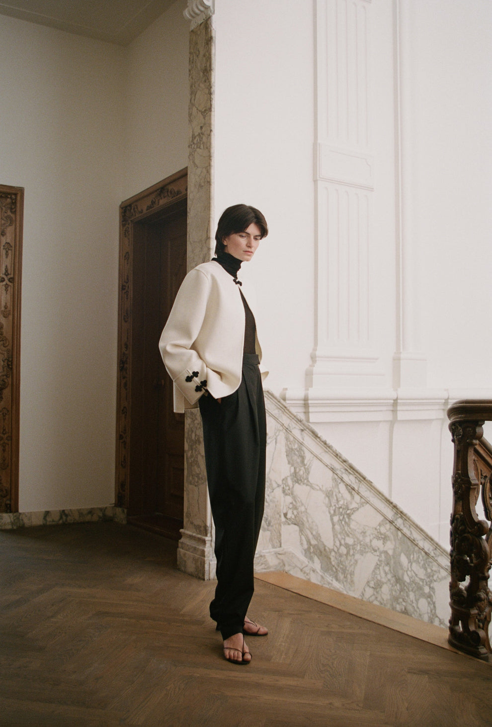 Woman standing in a room with white walls and wooden floor in a cream colored double faced wool jacket and black carrot shape trousers 