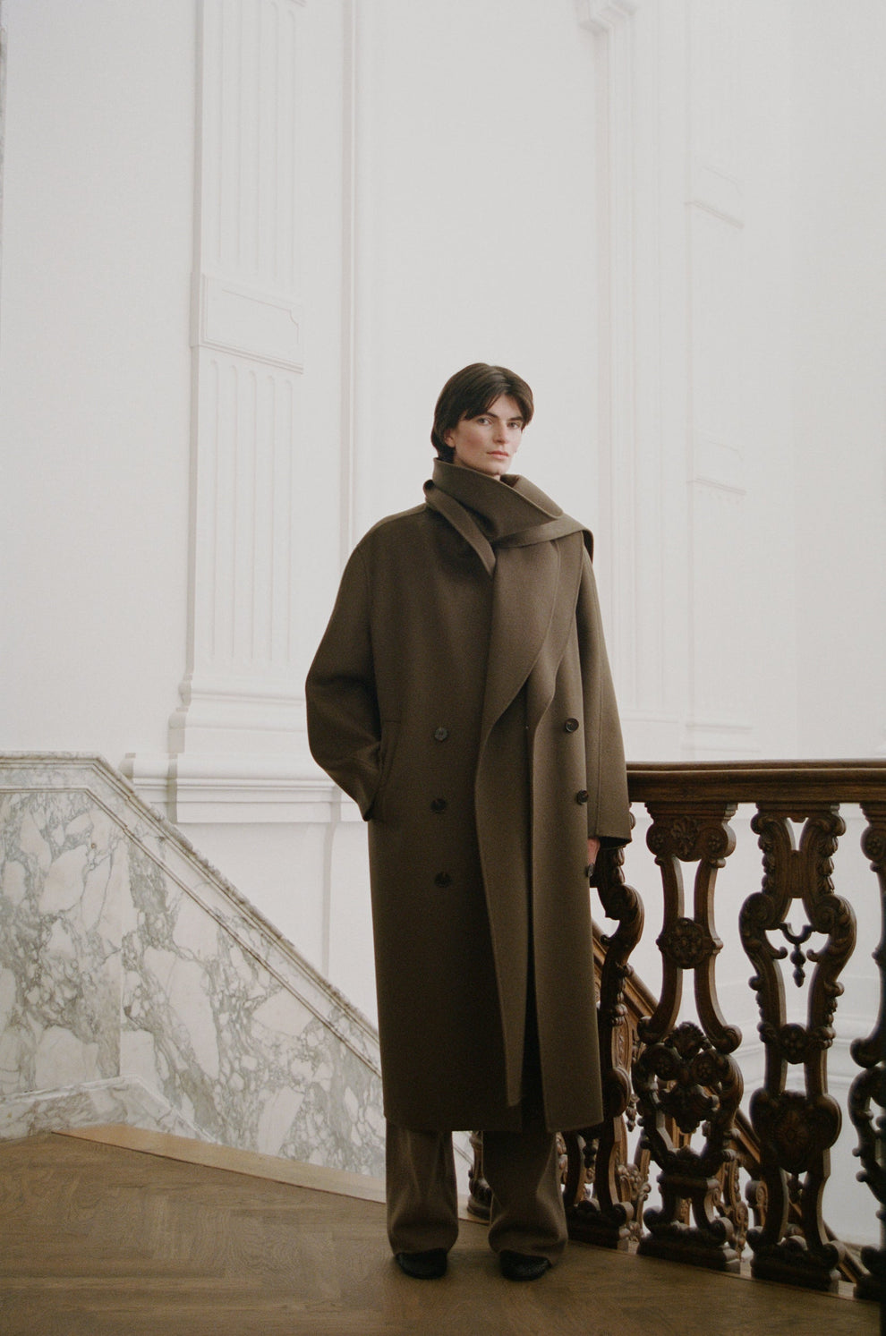 Person wearing a long taupe coat with scarf wrap detail standing in an elegant interior setting.