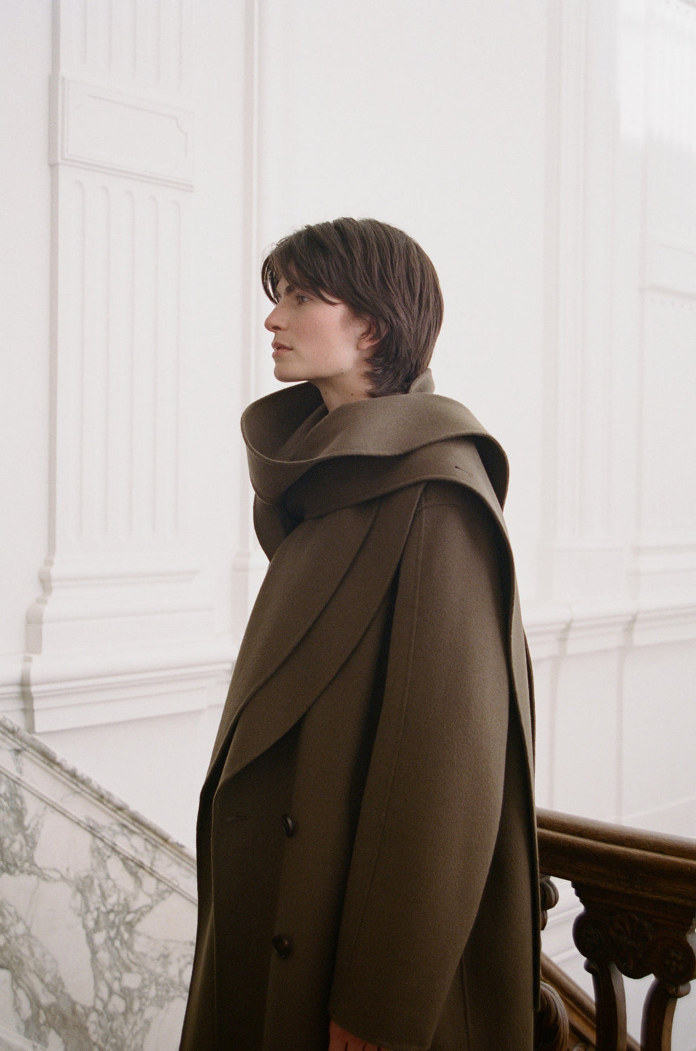 Woman wearing a taupe coat with a scarf wrap detail in an elegant interior setting