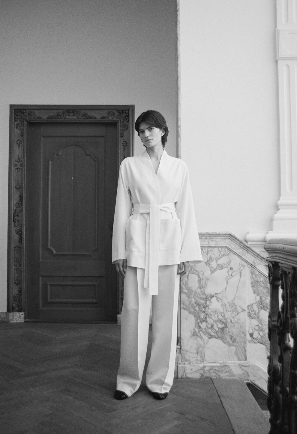 Black and white image of a woman wearing a kimono style cream colored short jacket and cream trousers standing in an elegant room