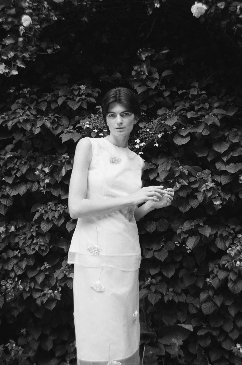 Woman in a white embellished sleeveless top with matching skirt standing against a leafy background