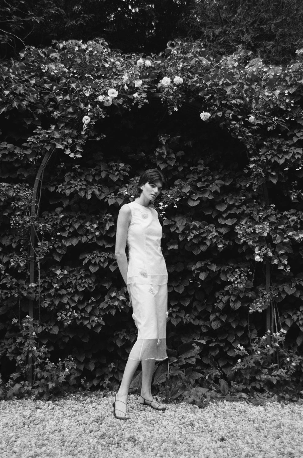 Woman in a white sleeveless embellished top and matching knee length skirt standing in front of a leafy background