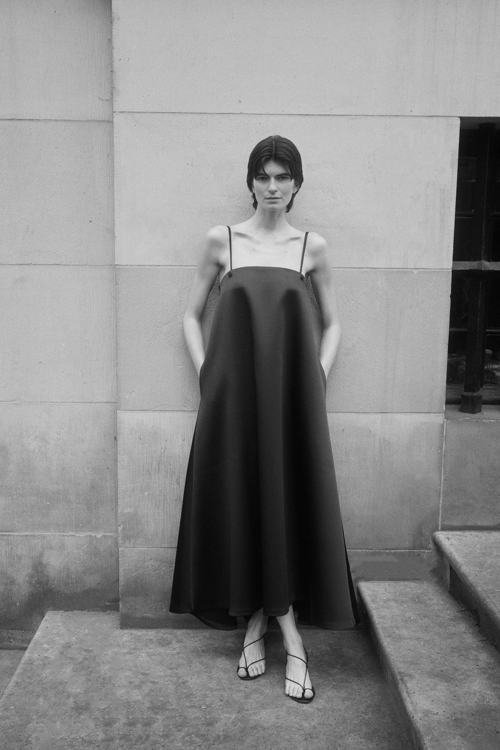 Woman wearing a black heavy satin a line dress with spaghetti straps standing against a concrete wall.