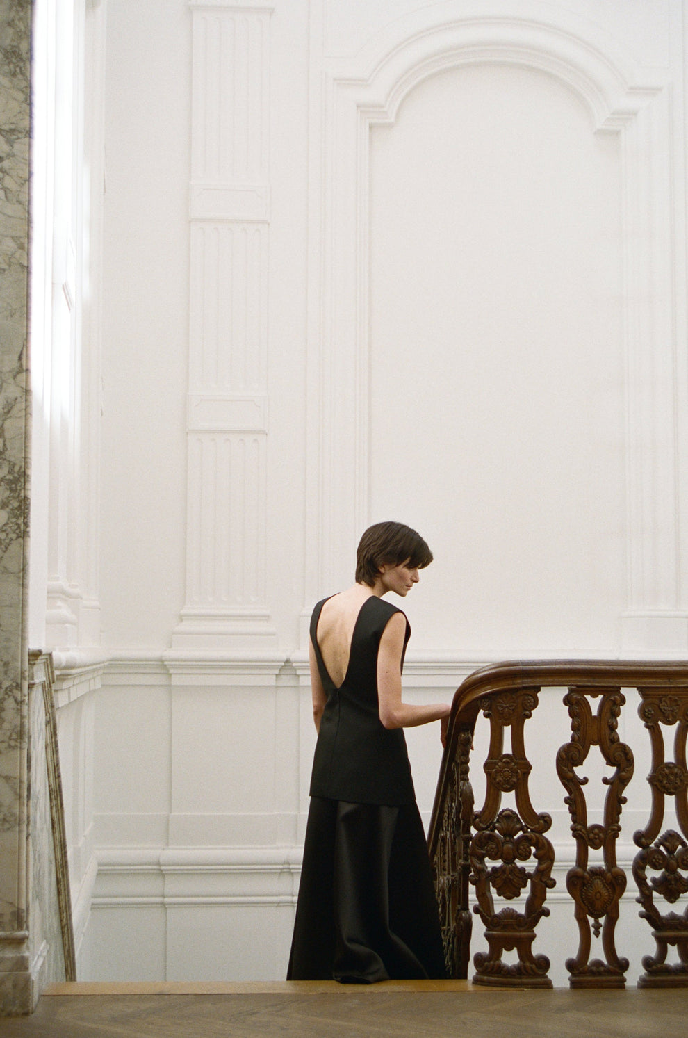 Woman in a black dress walking down elegant stairs with a wooden railing.