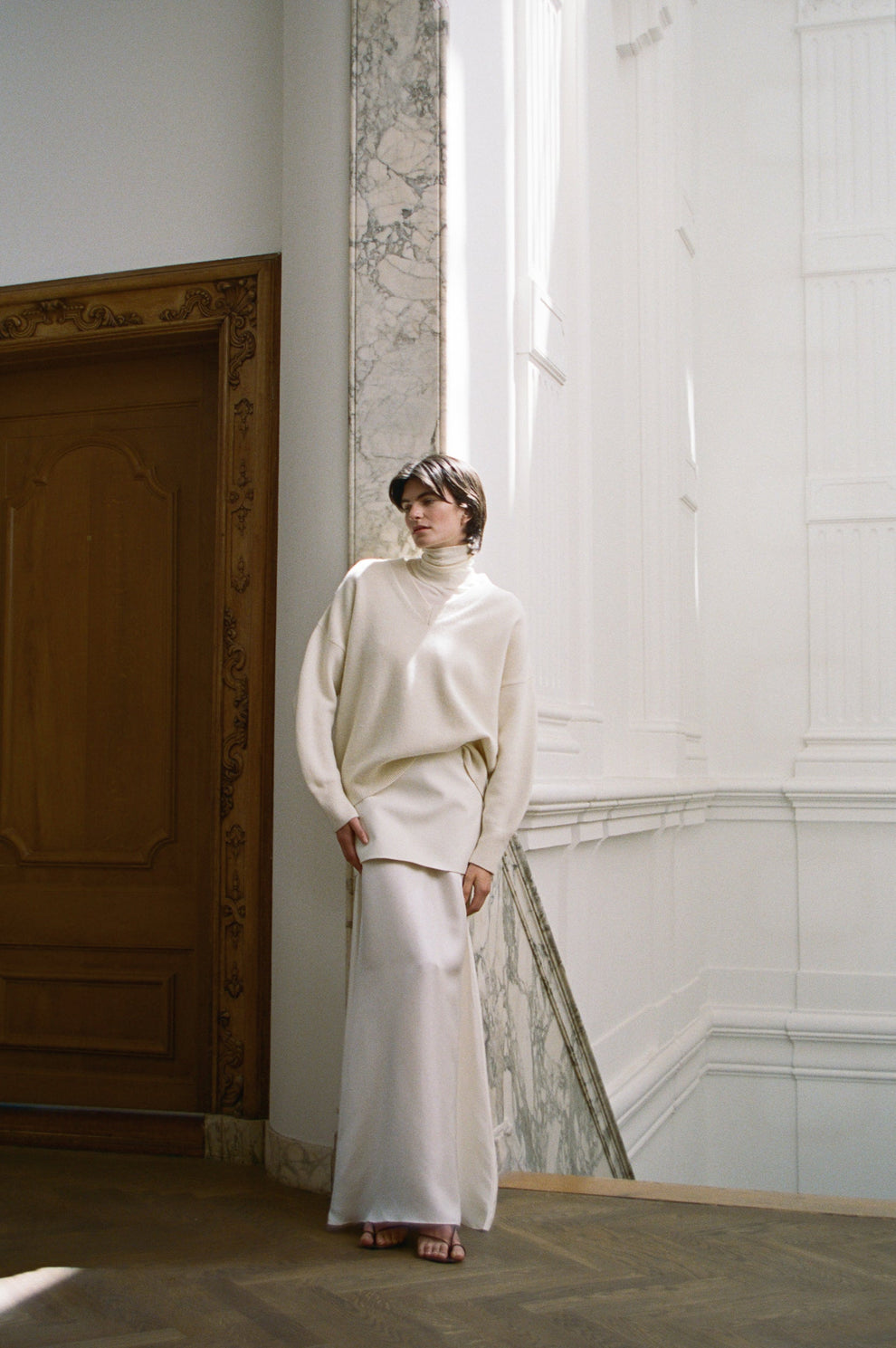 Woman in a cream v-neck sweater and floor length cream silk skirt standing in a room with a large door and decorative wall.