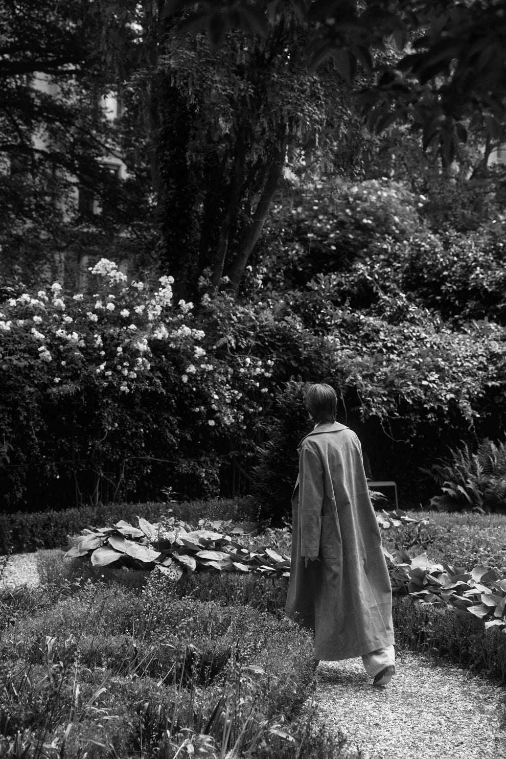 Woman walking in a suede trenchcoat through a garden with lush greenery and flowers