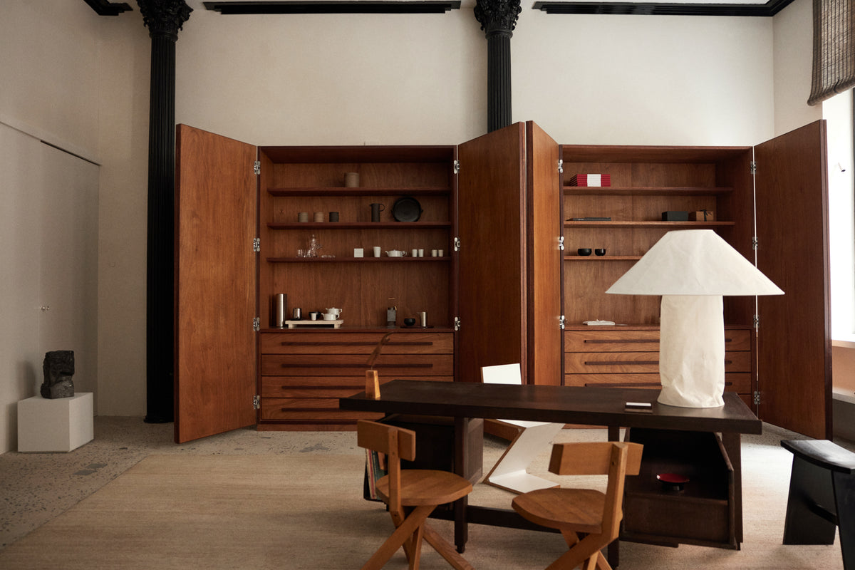 Historic interior with tall dark wood cabinetry, a long wooden table, midcentury chairs, a white table lamp, and black architectural columns.