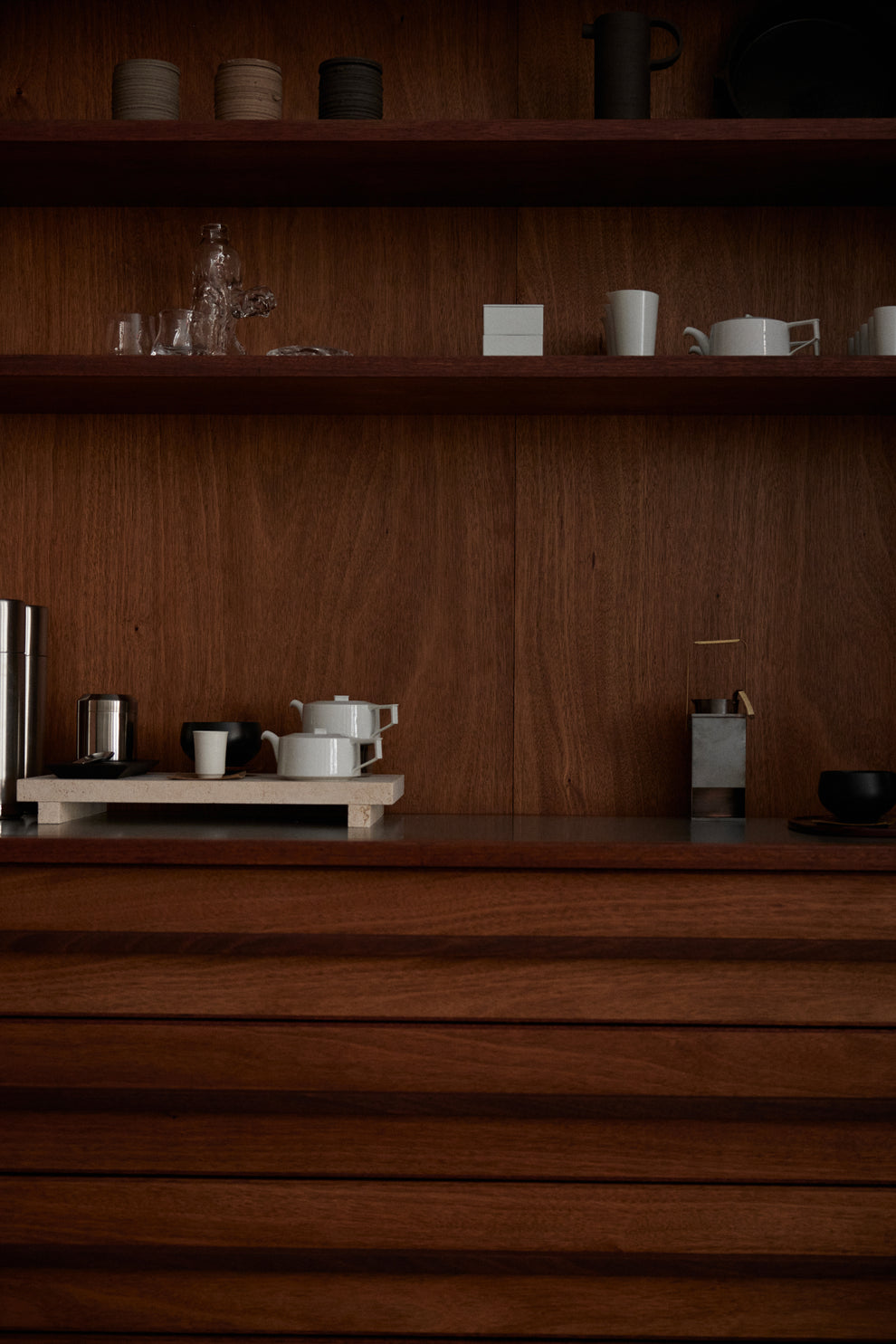 Dark wood shelving and cabinet displaying ceramic cups and bowls with a small coffee setup and a black kettle in a warm minimalist interior