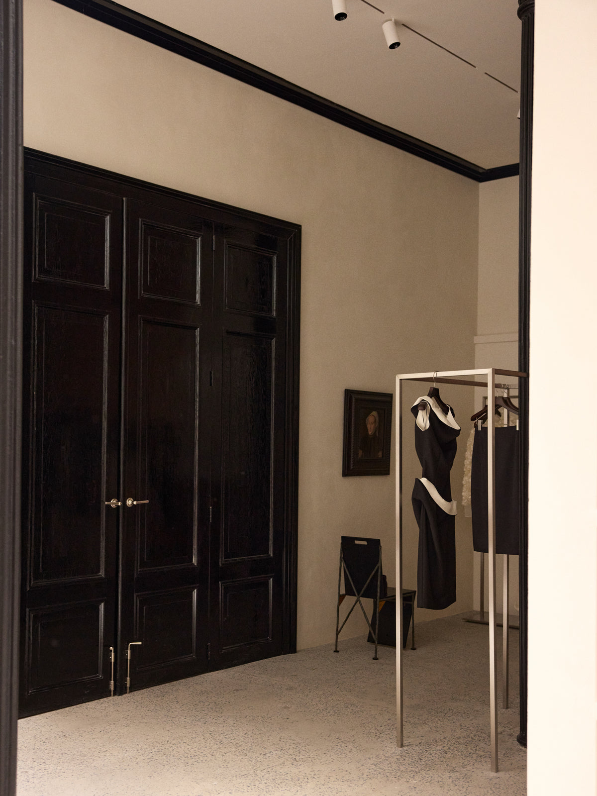 Elegant interior with dark paneled double doors, a freestanding clothing rack holding garments, a framed portrait, and a small chair against cream walls.