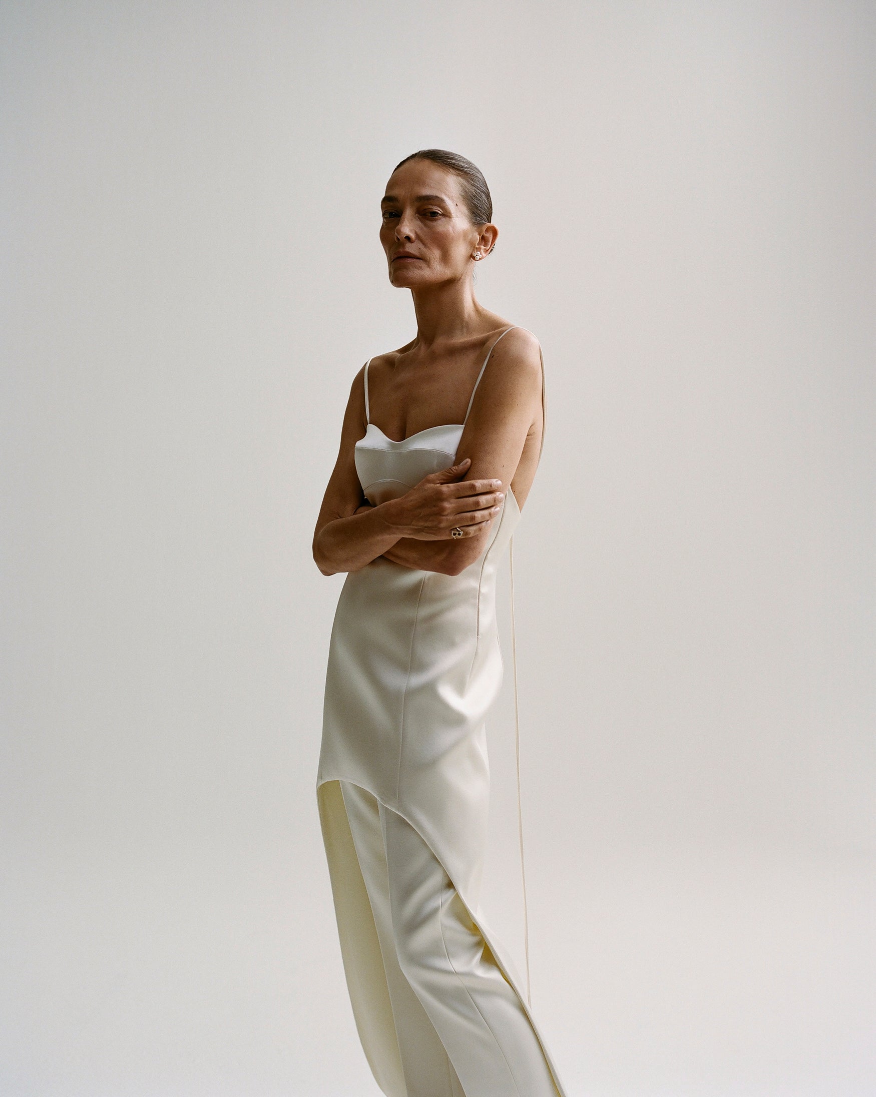 Woman in a satin long bustier dress with white pants underneath standing against a white backdrop.