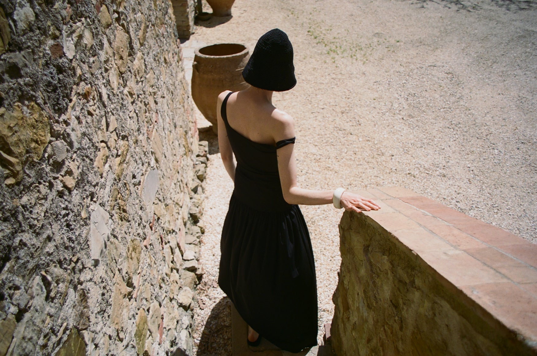 Woman in a black dress and black straw hat walking down stairs next to a stone wall and pavement.