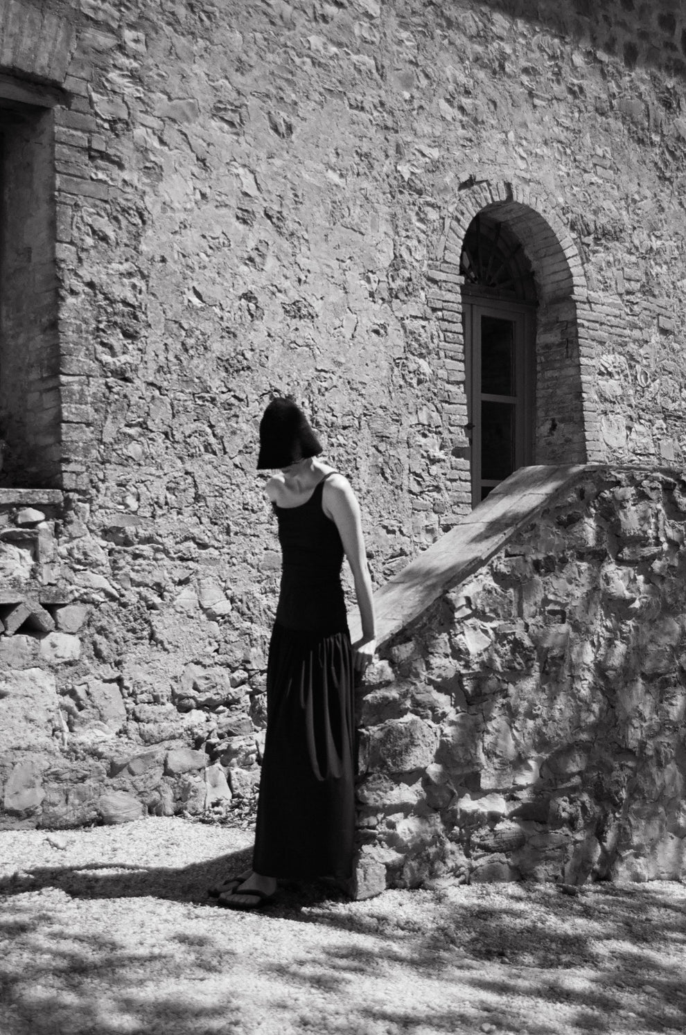 Woman in a long black dress and black straw hat standing against a stone wall