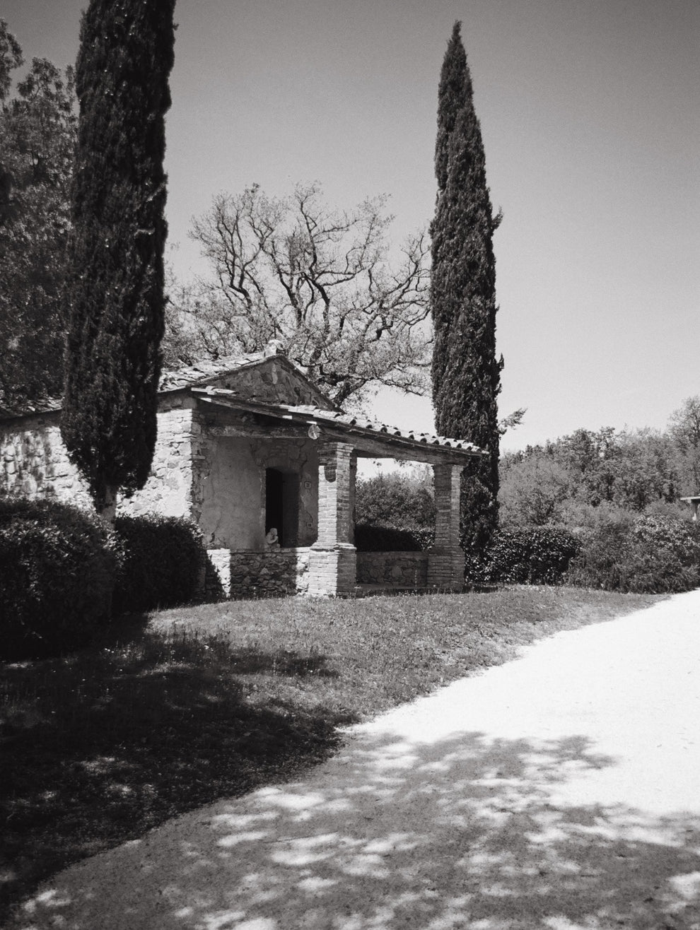 Black and white image of a rustic stone building with cypress trees in a garden setting