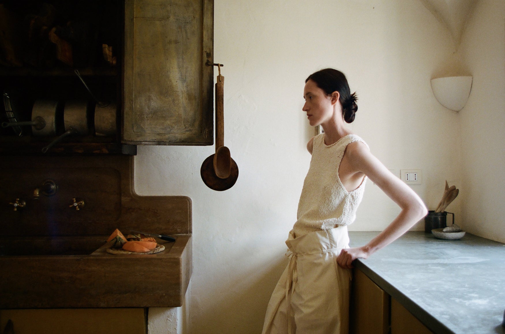 Woman standing in a kitchen next to a stove in a sleeveless cream boucle top and beige cotton trousers
