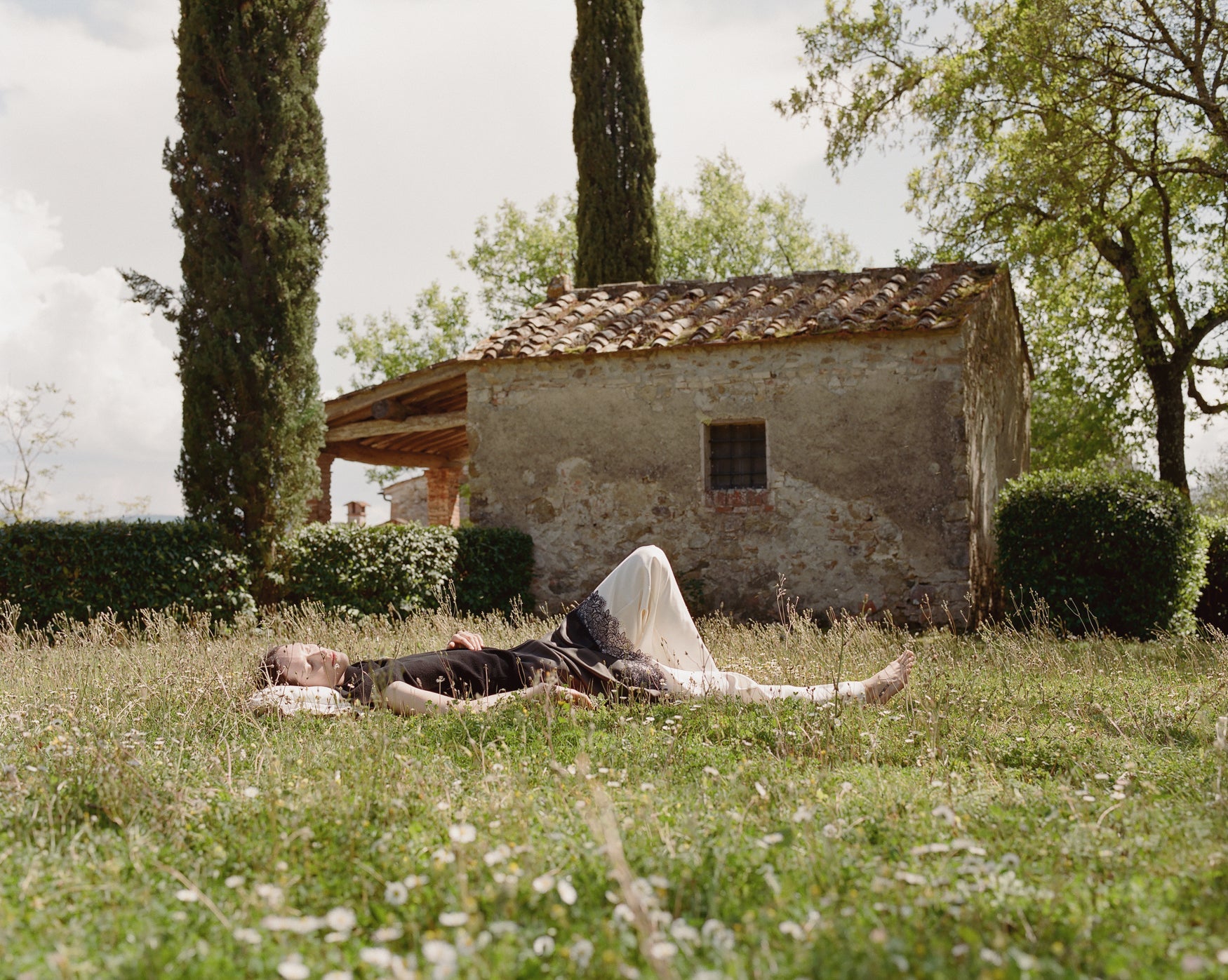 Woman lying on grass in front of a rustic building with trees in the background wearing cream trousers and a lace trimmed skirt and black top
