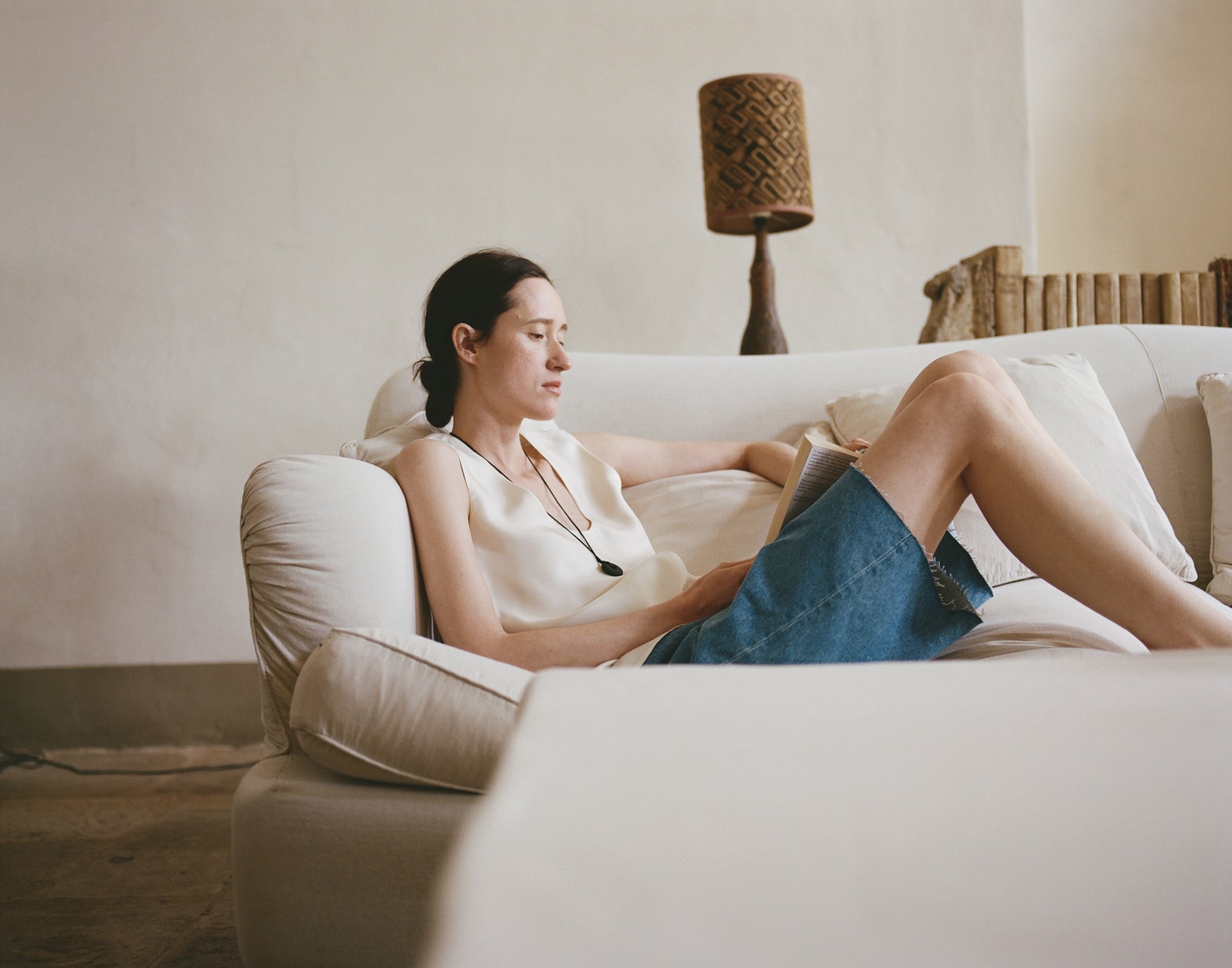 Woman sitting on a couch reading a book in a cozy living room wearing a cream silk top and baggy denim shorts