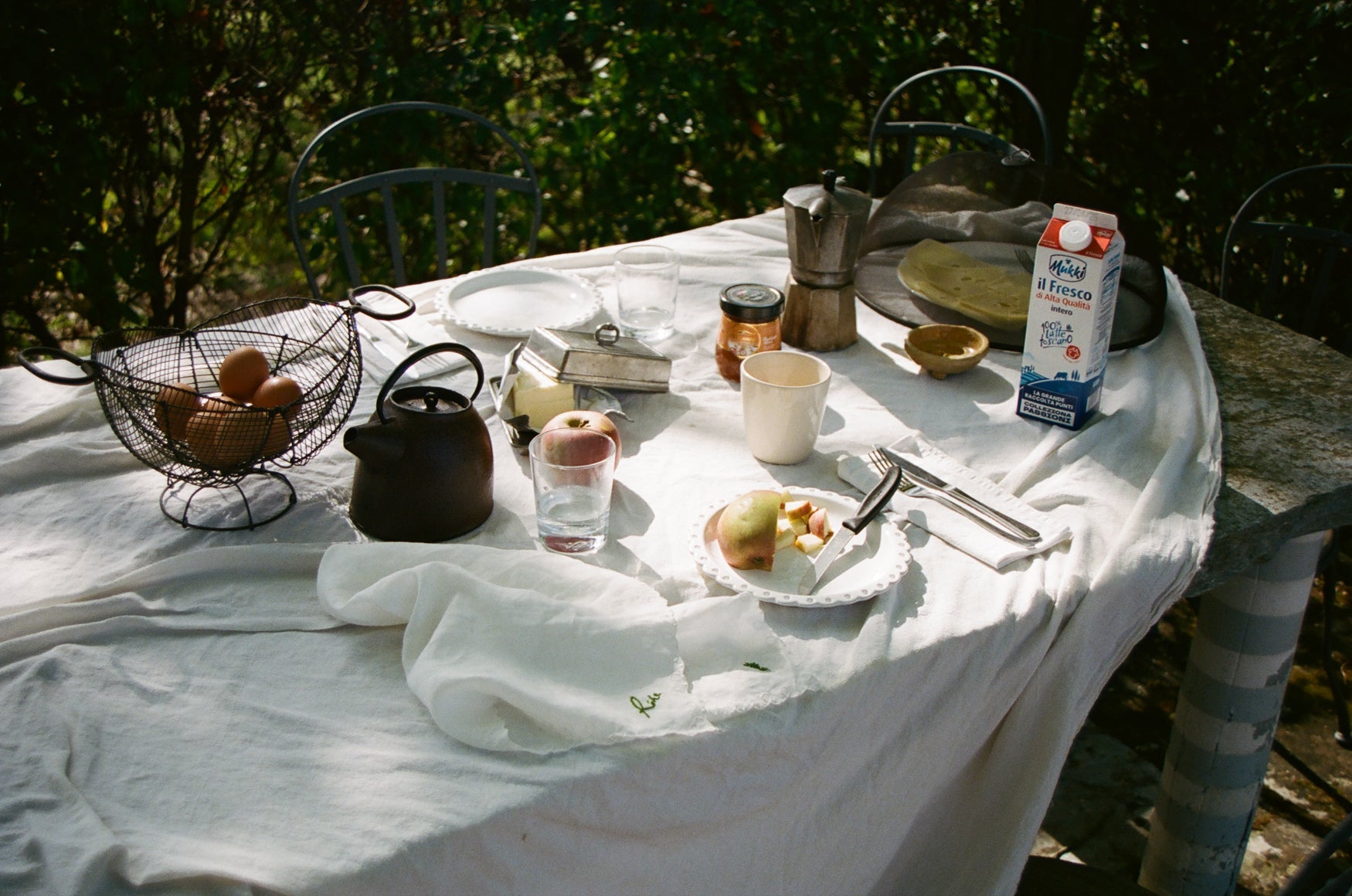 Table set with breakfast items including a teapot, fruit, and milk carton outdoors.