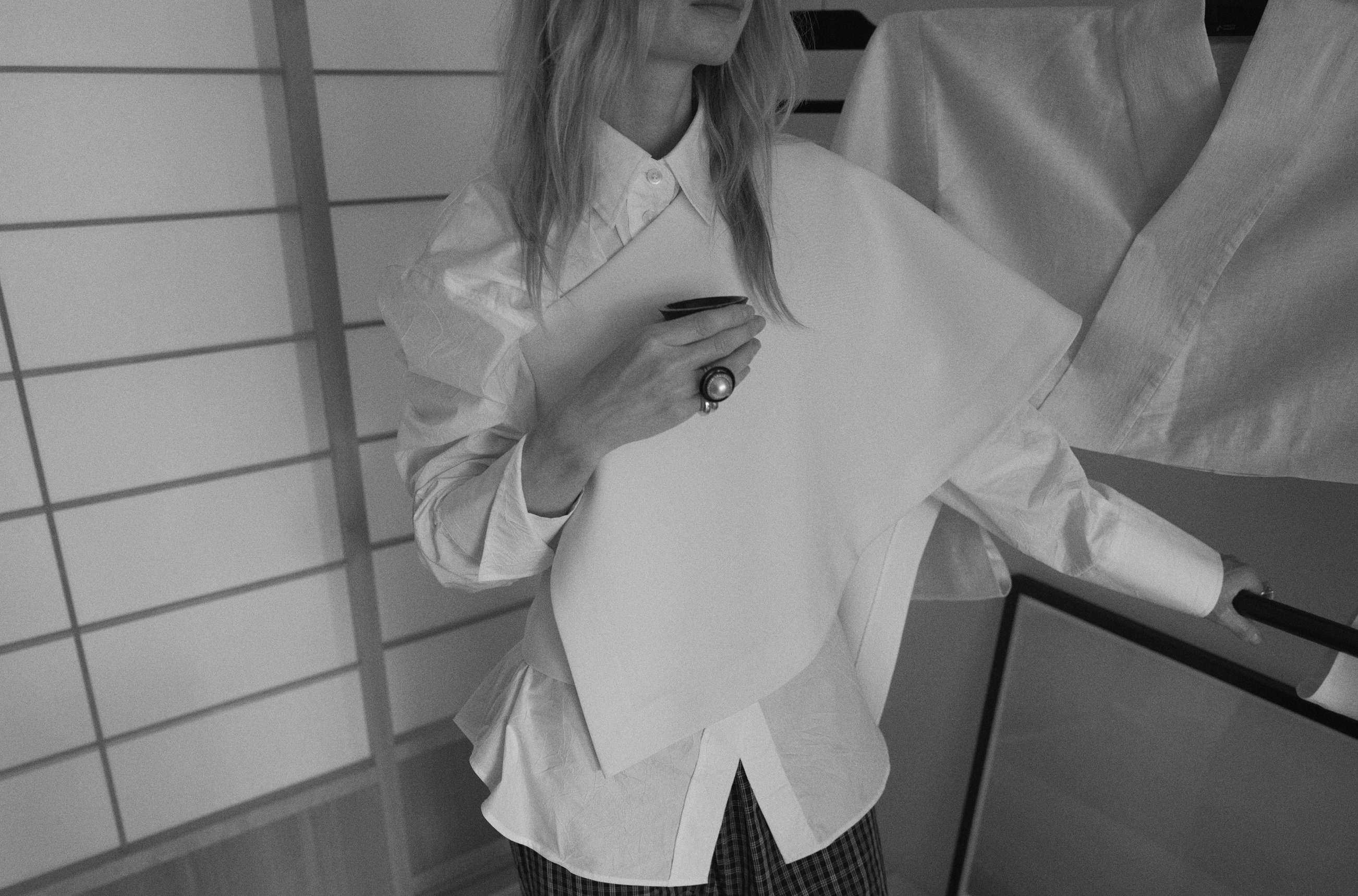 Close-up image of a woman wearing an asymmetrical top layered over a white shirt, standing in a Japanese interior