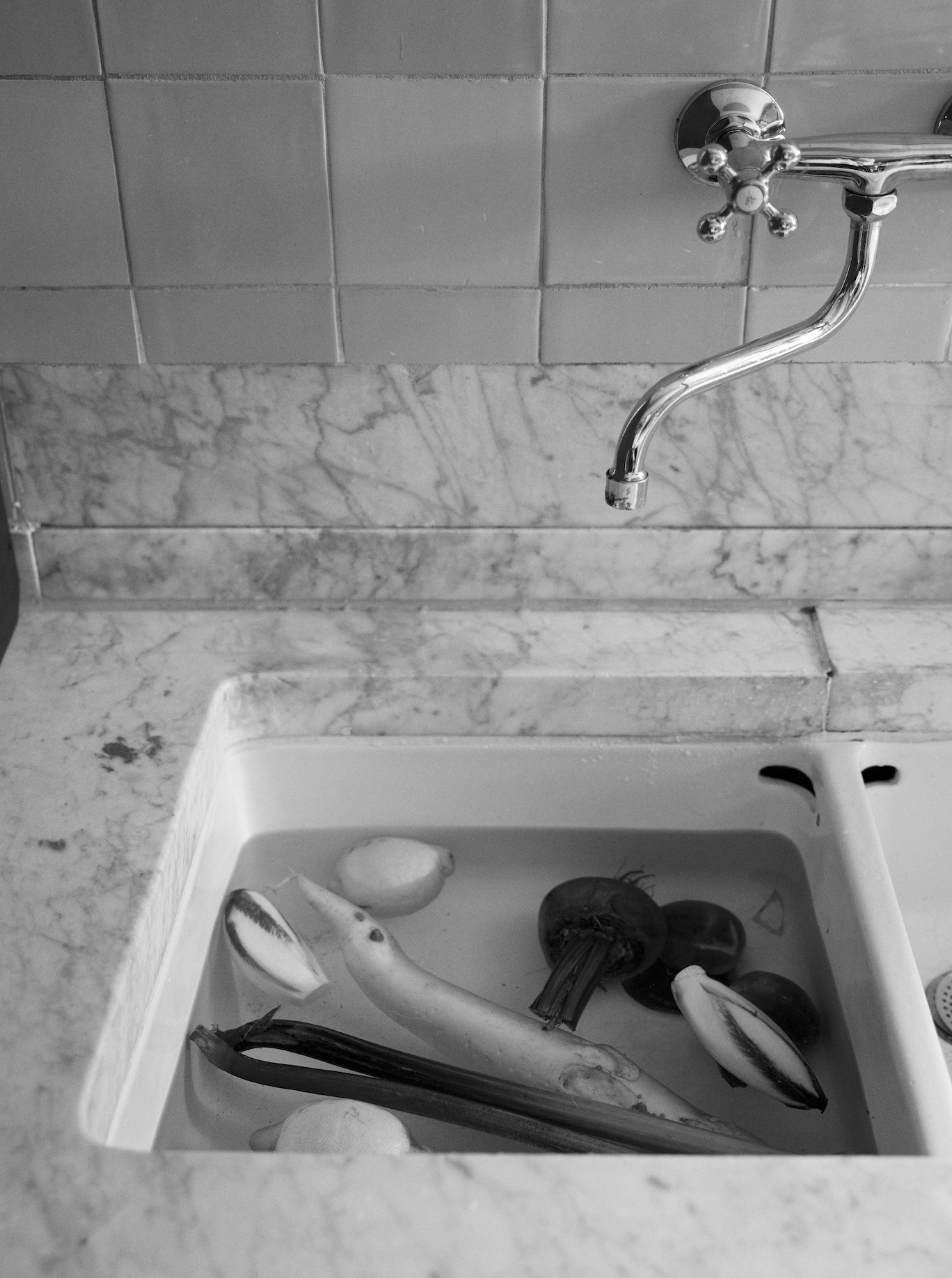 Black and white image of a marble kitchen sink with various vegetables soaking in water under a chrome faucet, featuring the product Róhe Cookbook | red.
