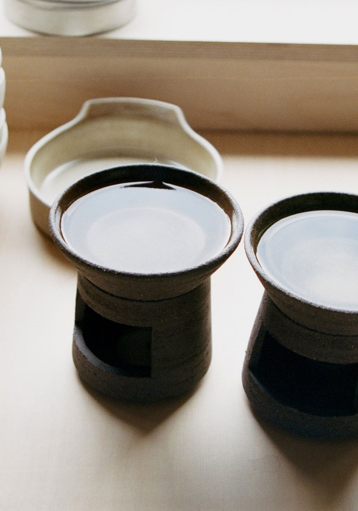 Two dark brown ceramic oil burners filled with water on a light surface with a light ceramic dish in the background, showcasing the Ceramic Oil Burner | soil design.