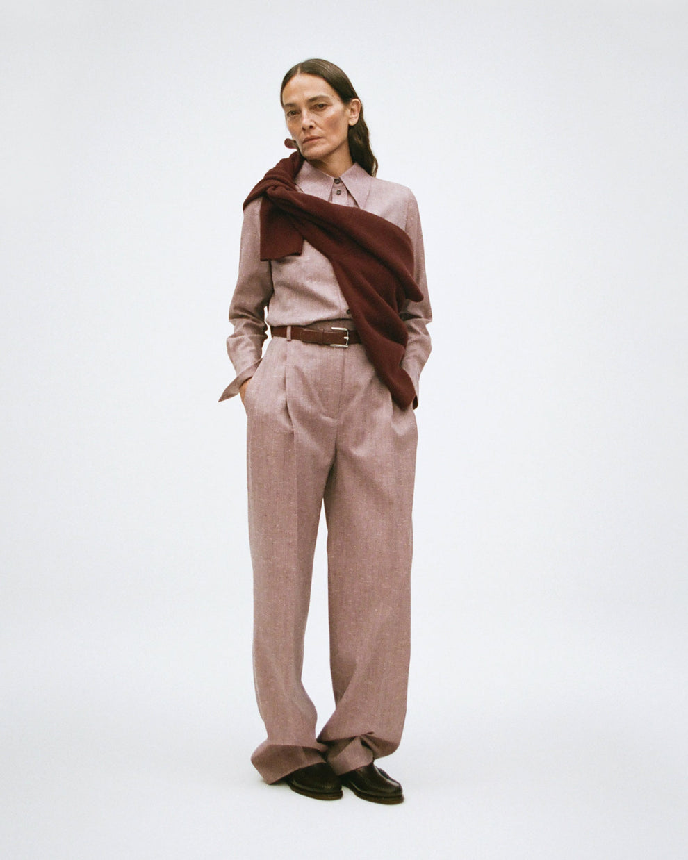 Woman wearing a pink tweed shirt with matching trousers with a belt and burgundy sweater tied around the shoulders on a white background