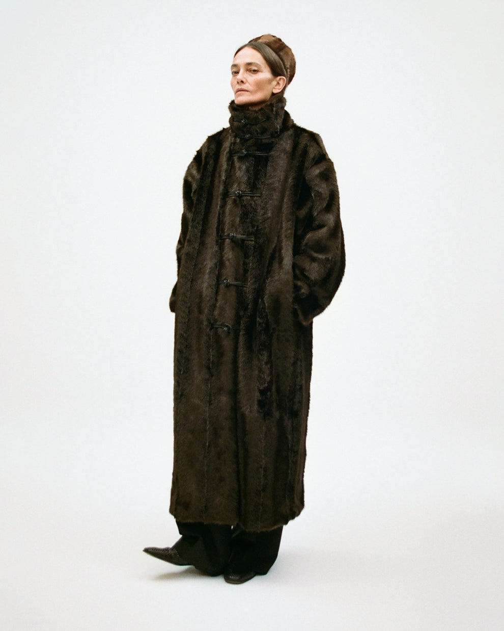 Woman wearing a long dark brown fake fur coat and matching pillbox hat on a white background