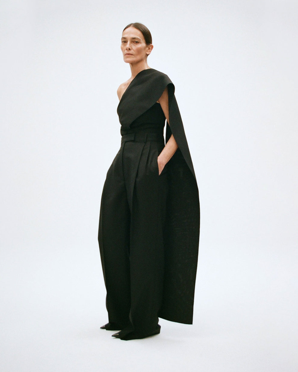 Woman wearing a black one-shoulder top with a cape with black wide leg pants on a white background